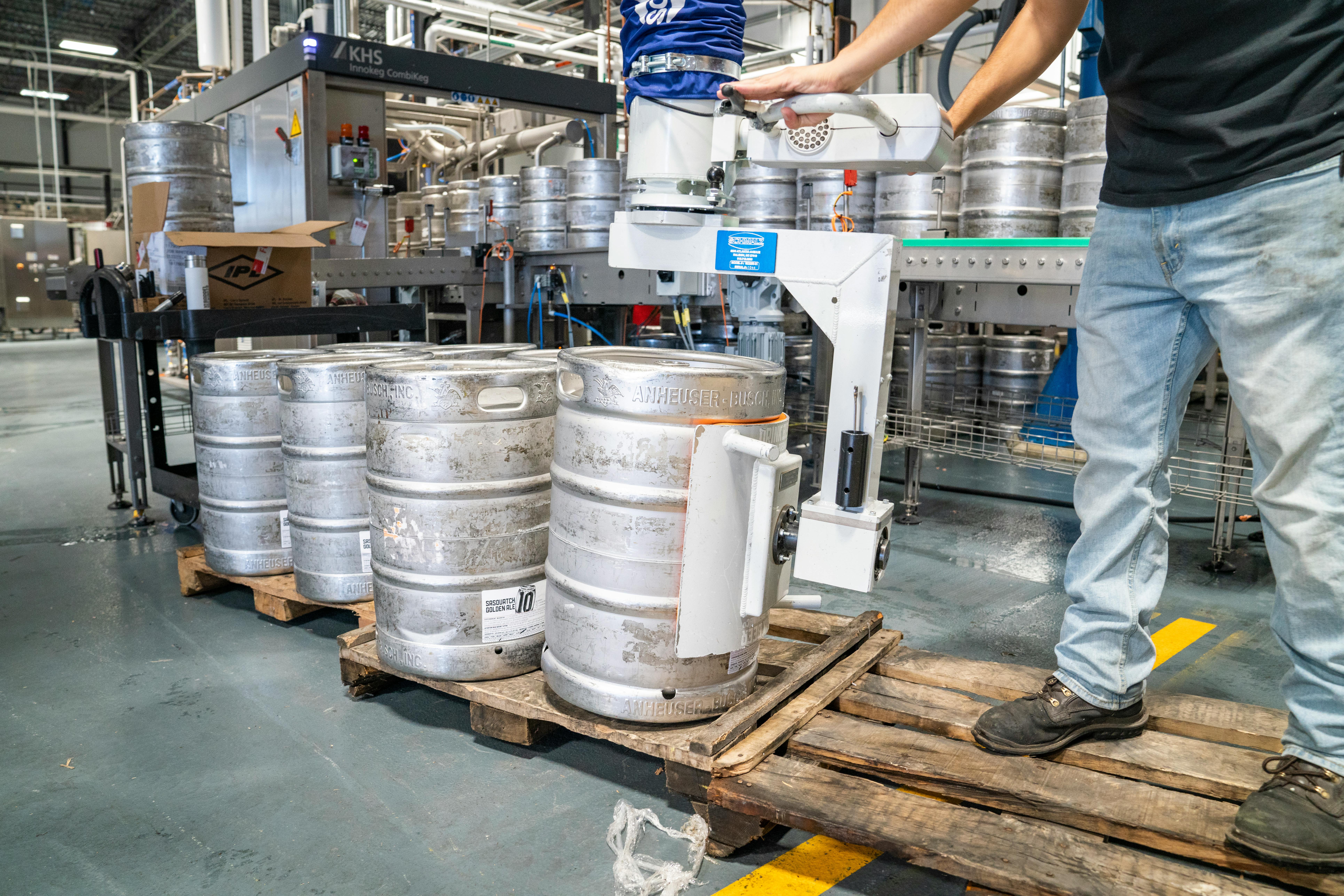 A worker operates machinery to stack steel kegs in a brewery warehouse.